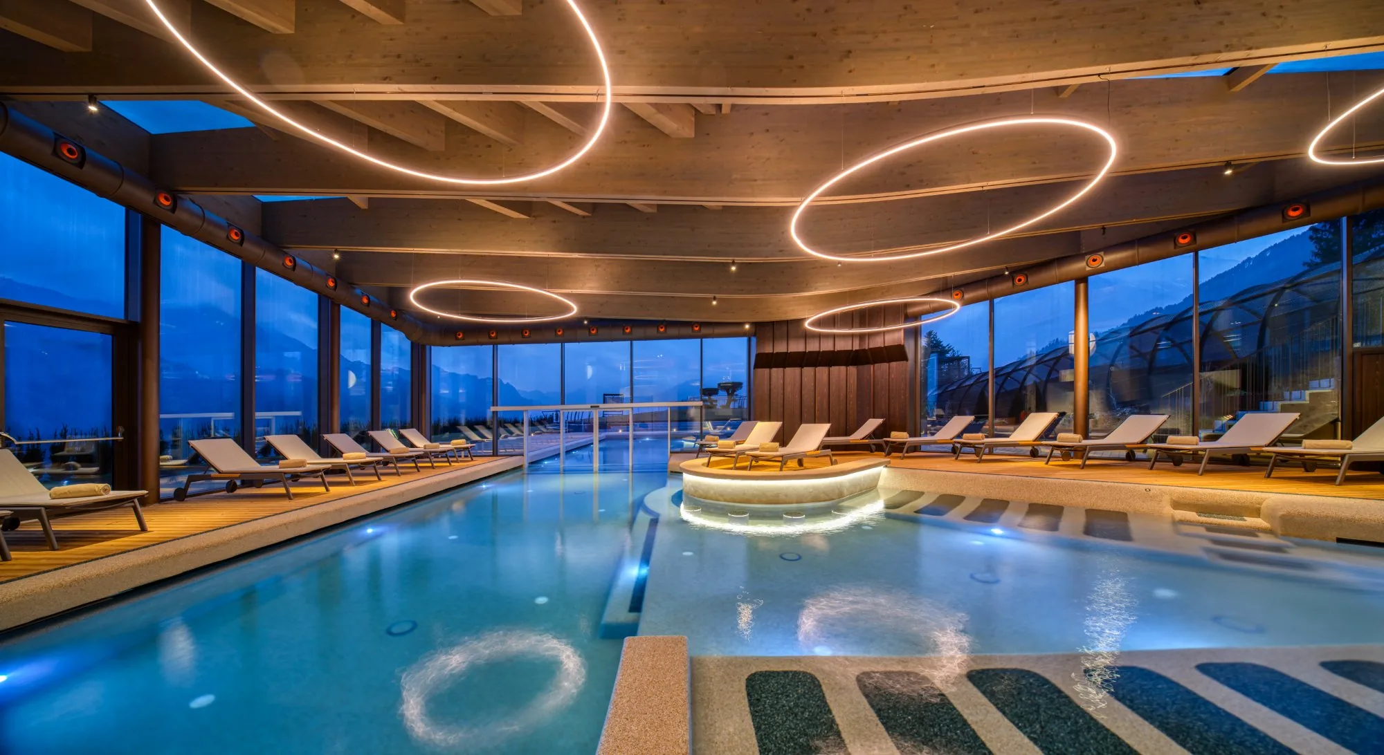 Piscina interna riscaldata con lettini, soffitto in legno con lampadari ad anello, vetrate panoramiche sul paesaggio serale della Valseriana nella My Purity SPA di Clusone