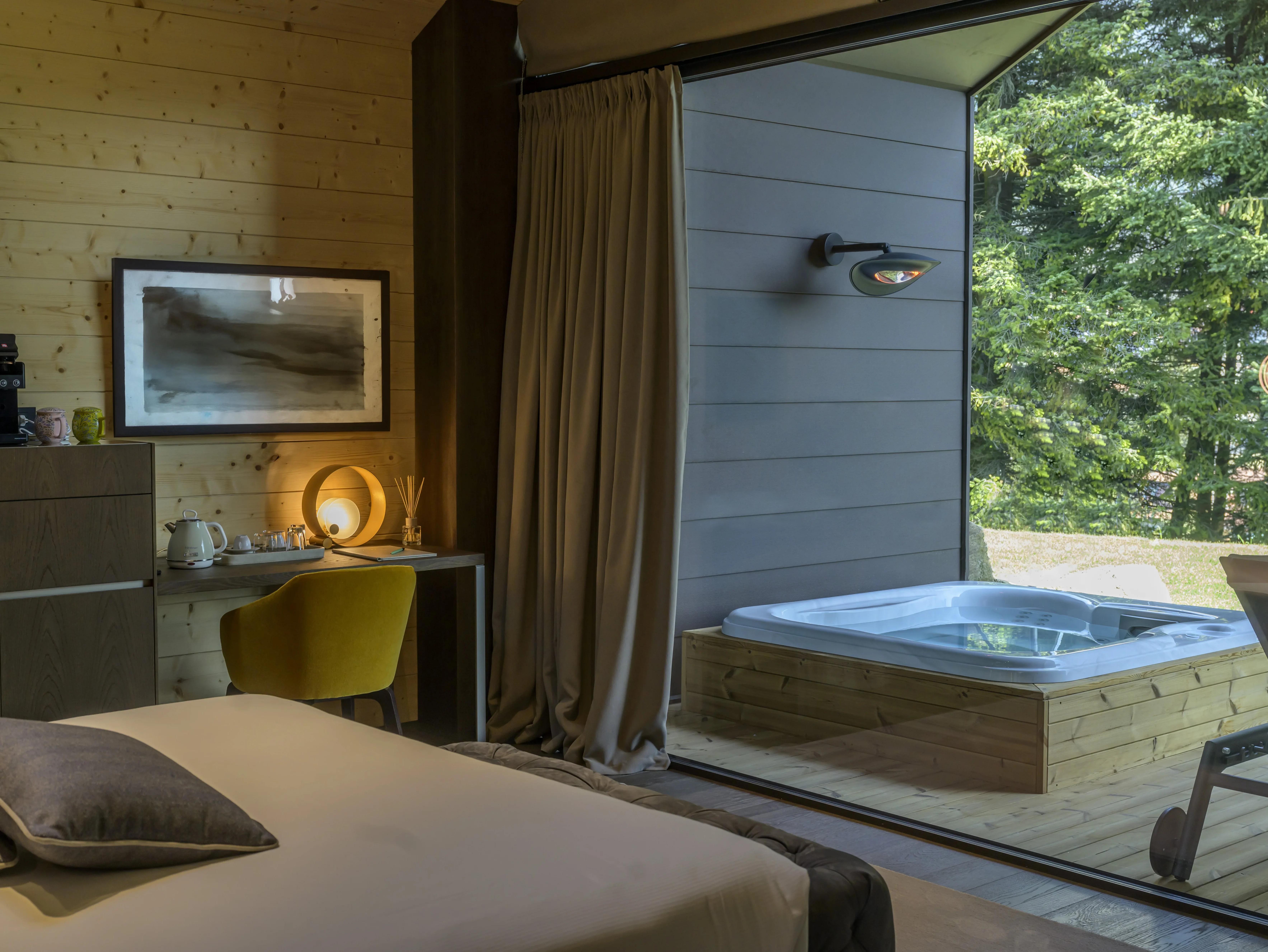 Camera chalet con letto, scrivania, sedia gialla e vetrata aperta su terrazza con idromassaggio privato e abeti