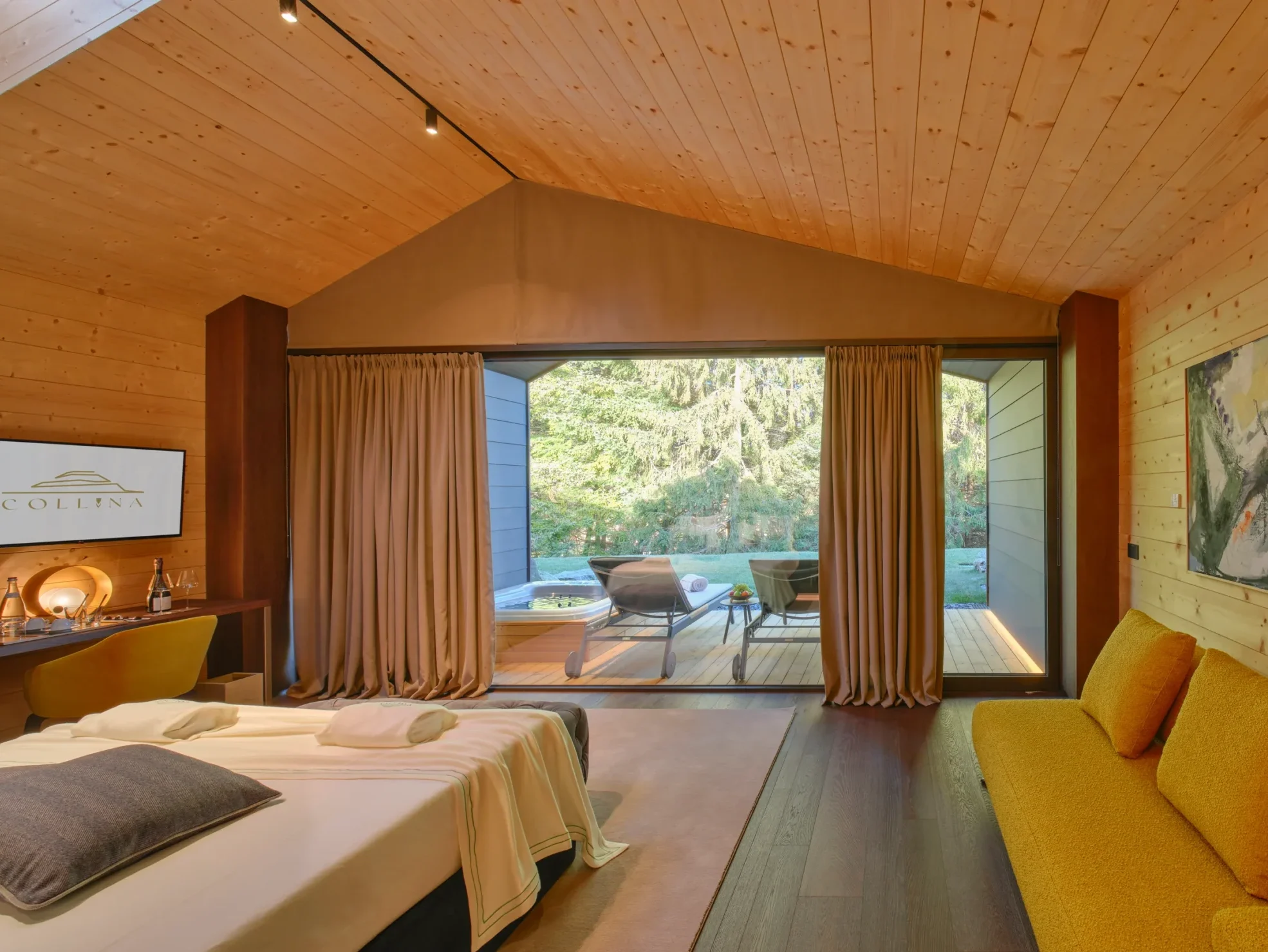 Camera chalet con letto king-size, divano giallo, TV con logo Collina e vetrata aperta sulla terrazza con giardino
