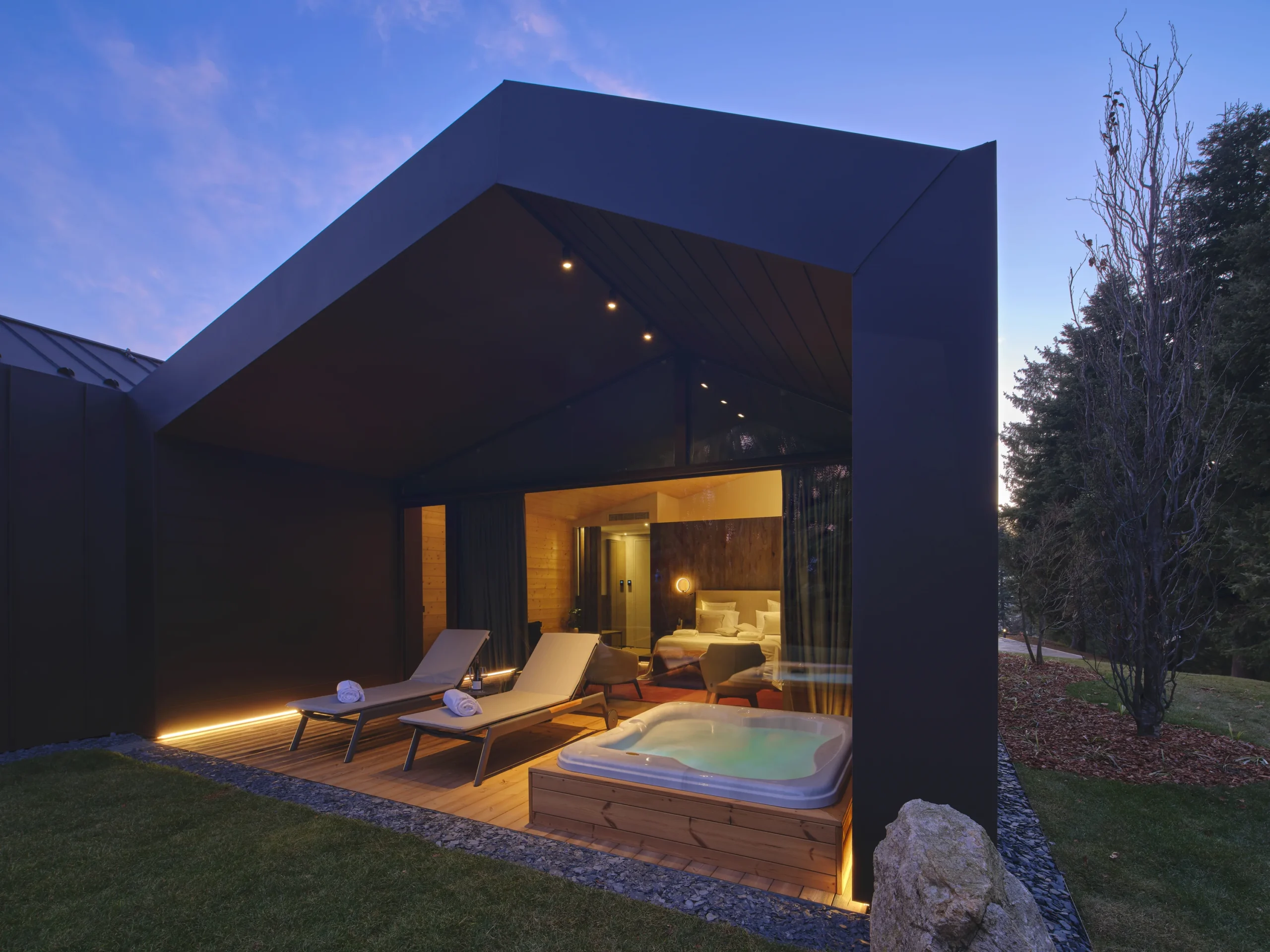 Chalet con struttura scura, terrazza in legno illuminata, jacuzzi privata e lettini di notte con camera visibile all'interno