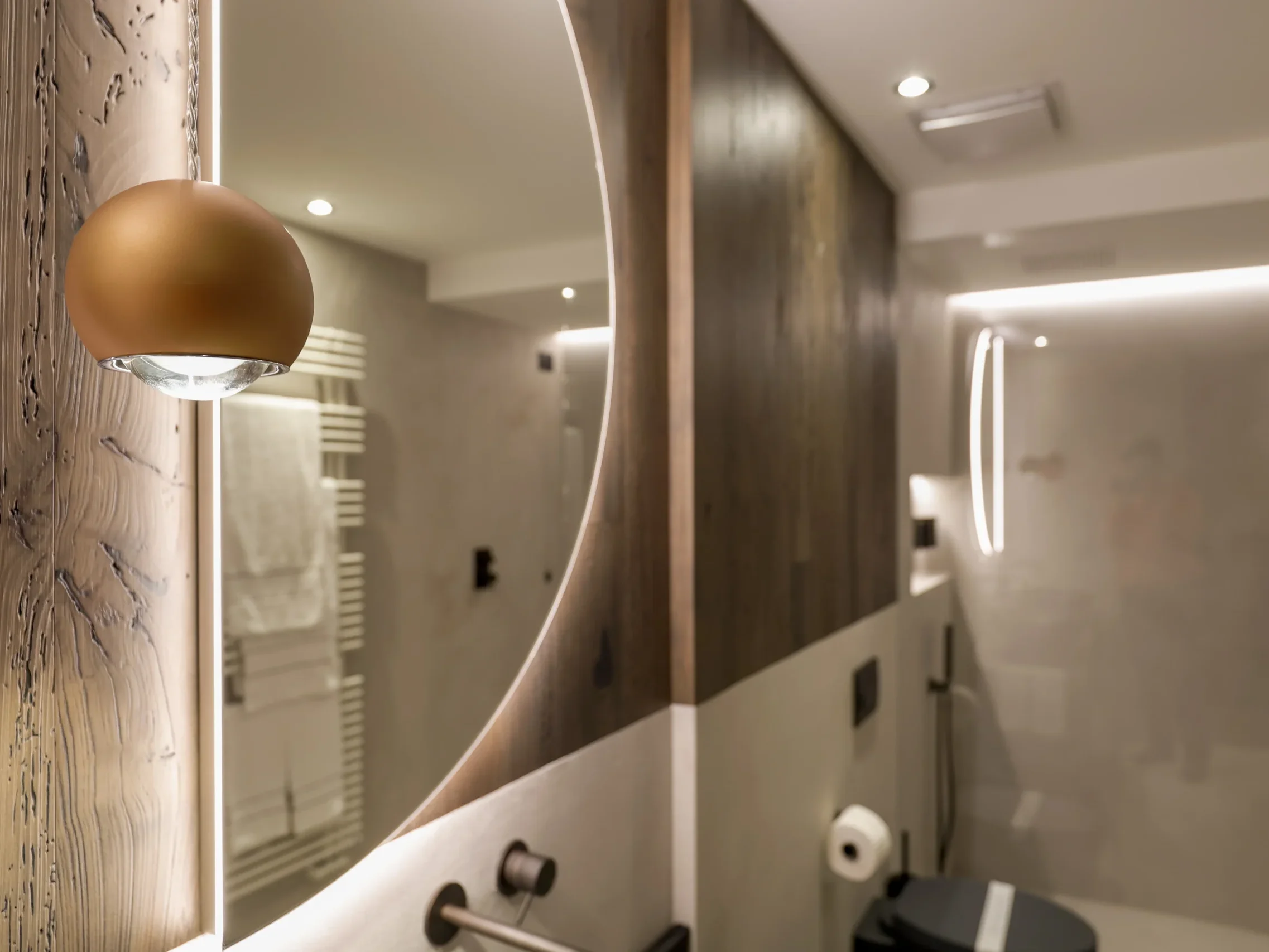 Dettaglio bagno chalet con specchio tondo retroilluminato, applique sferica dorata e parete in legno antico
