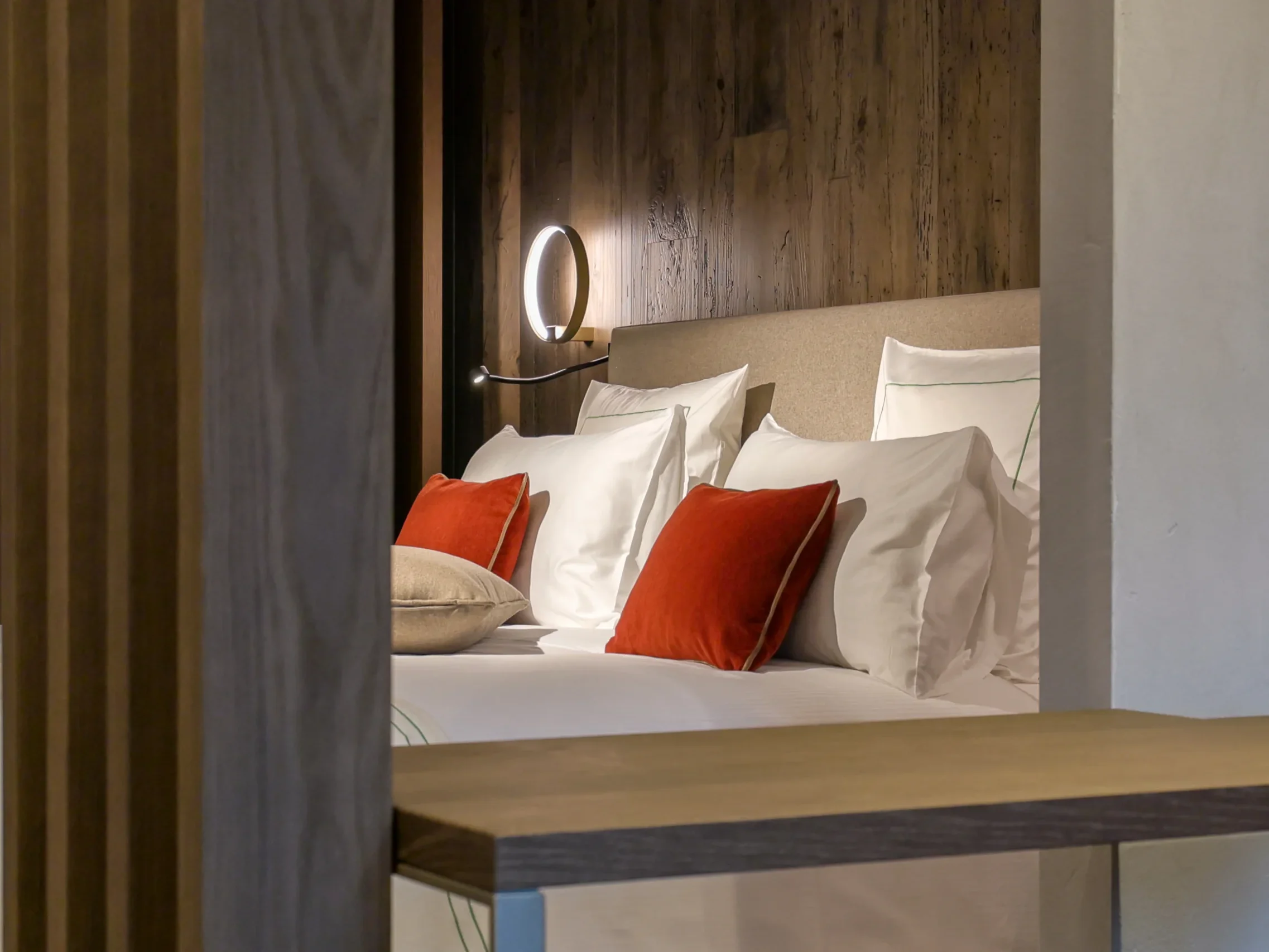 Dettaglio letto con cuscini bianchi e rossi, testata imbottita, applique circolare illuminata e parete in legno antico scuro