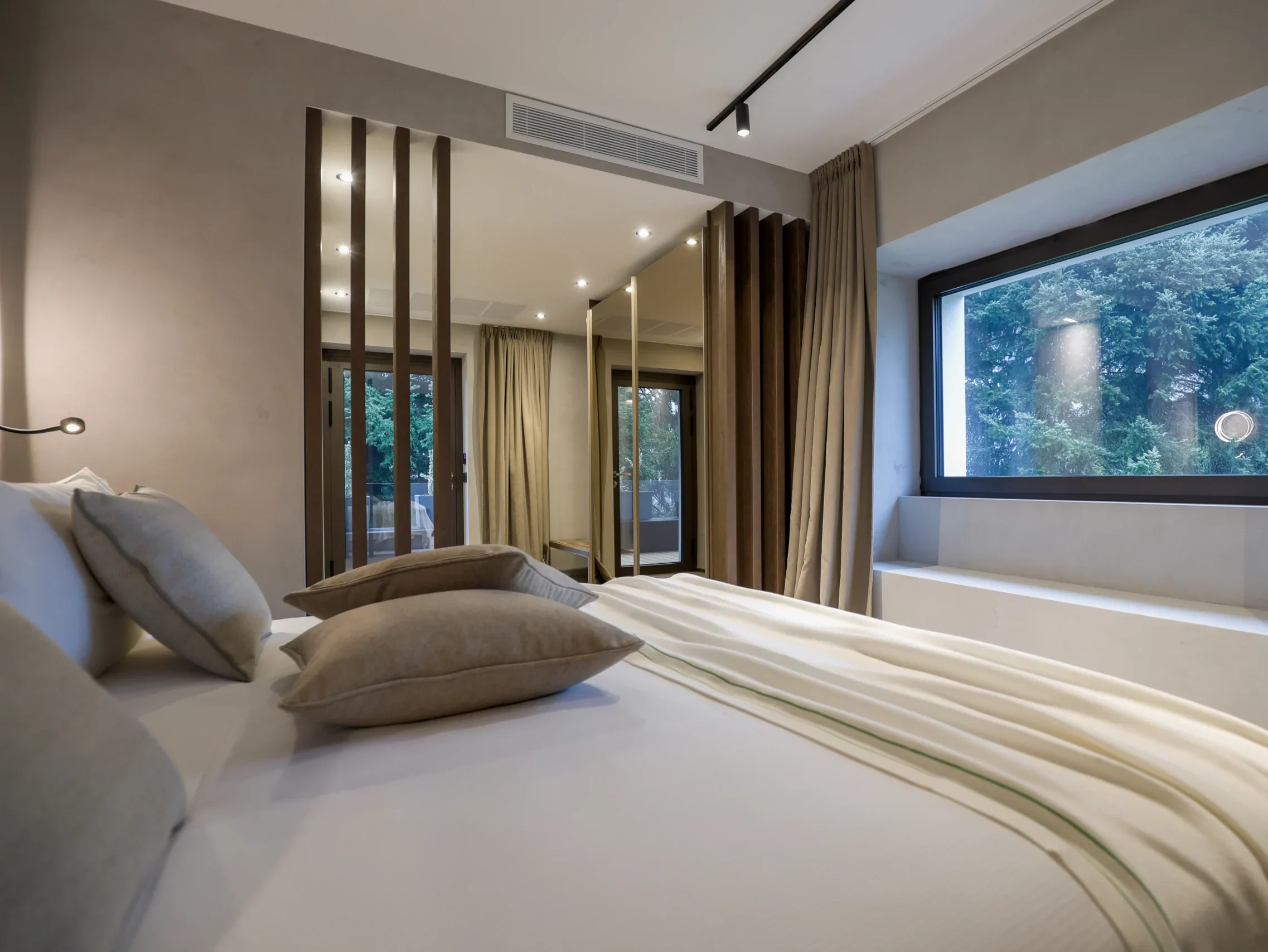 Camera Junior Suite con letto, cuscini grigi, divisori in legno, armadio specchio e finestra panoramica sul giardino