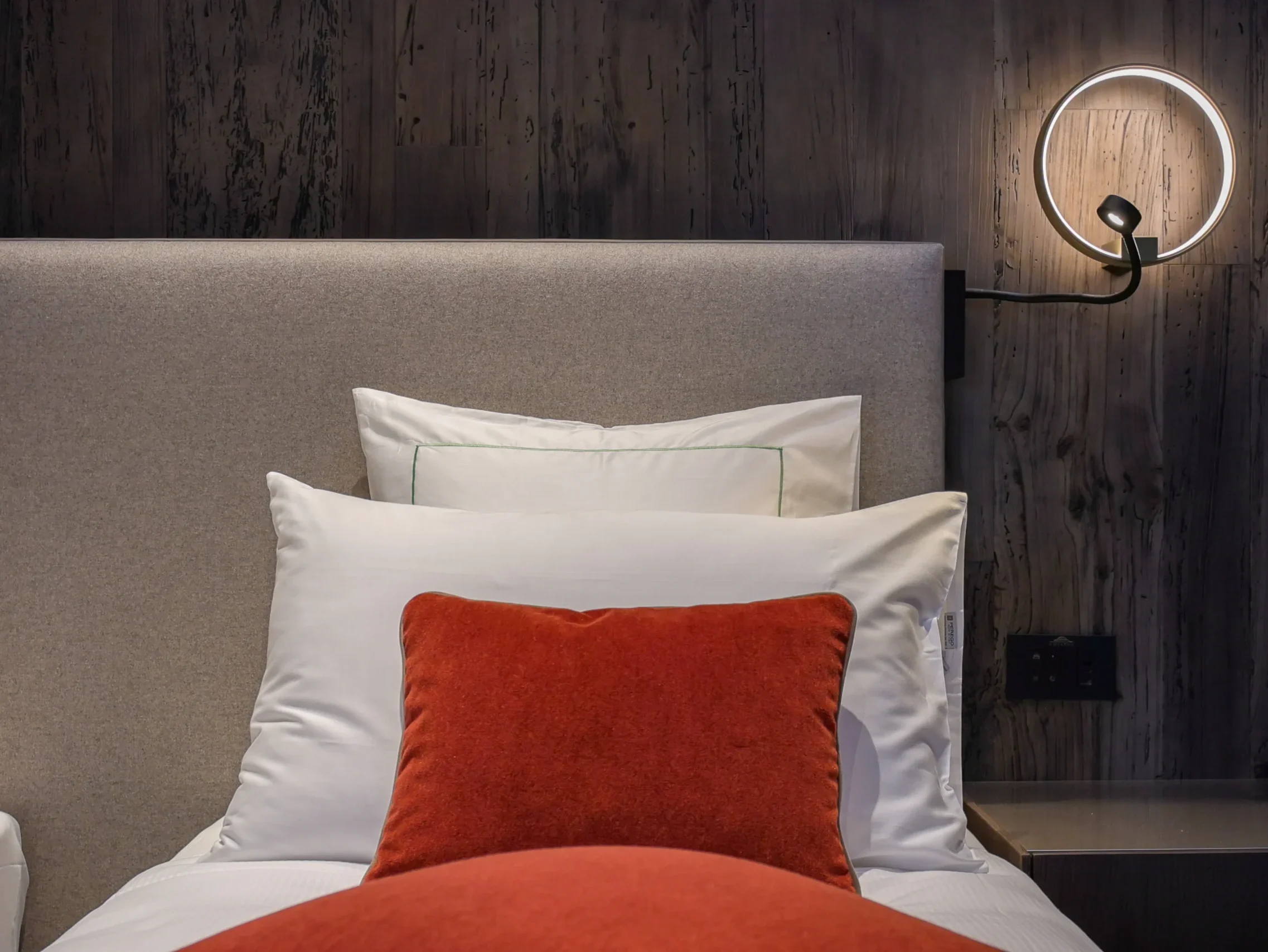 Testata letto imbottita con cuscini bianchi e cuscino rosso mattone, applique circolare illuminata e parete in legno antico scuro
